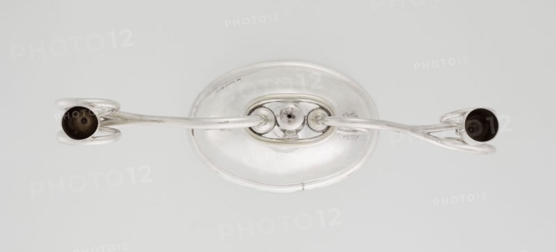 Two-candle candlestick by William Leuchars fl. 1876–1888 and Leuchars & Son goldsmiths, fl. c.1845–1902. Two-branched candlestick on a circular, domed foot; the shaft rests on a ball no finial, enclosed at three-quarters of its height in a plate-like ring, from which extend movable arms made of S-shaped twisted wires, split at the ends, terminating in cylindrical candle sockets. The foot is engraved with the Pilawa coat of arms with the motto beneath a nine-baton count's crown. Inscription/provenance notes: Pilawa coat of arms, Potocki coat of arms, rank crowns, mottos; scrap – purchase, purchase. Date: 1881/1882. Place of origin: London, England, Great Britain. Materials/function: lighting. Dimensions: height 27 cm, width 32 cm, depth 11.5 cm; weight 1082 g., A polished metallic object with a rounded base and two curved arms extending outward to hold a pair of cup-shaped ends; the metal is primarily silver with darker inner surfaces visible inside the cup-like ends, and the background...