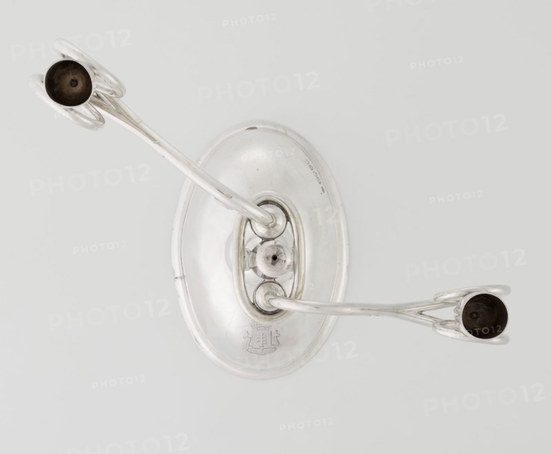 Two-candle candlestick, maker: William Leuchars fl. 1876–1888; firm: Leuchars & Son London; goldsmith's firm; fl. ca. 1845–1902. Silver candlestick with two movable arms made of S-shaped twisted wires, split at the ends, terminating in cylindrical candle sockets. Two-branched candlestick on a circular domed foot; shaft rests on a ball no finial and is enclosed at three-quarters of its height in a plate-like ring from which the arms extend. Foot engraved with the Pilawa coat of arms beneath a nine-baton count's crown and motto. Date: 1881–1882. Place of origin: London, England, Great Britain. Function: lighting. Materials/technique: goldsmithing/silver. Dimensions: height 27 cm; width 32 cm; depth 11.5 cm; weight 1082 g., A metallic wall-mounted fixture with an oval backplate and two curved arms extending outward, each arm ending in a shallow cup; the object is shiny silver with some darker interior areas in the cups and slight surface blemishes, set against a pale gray background.