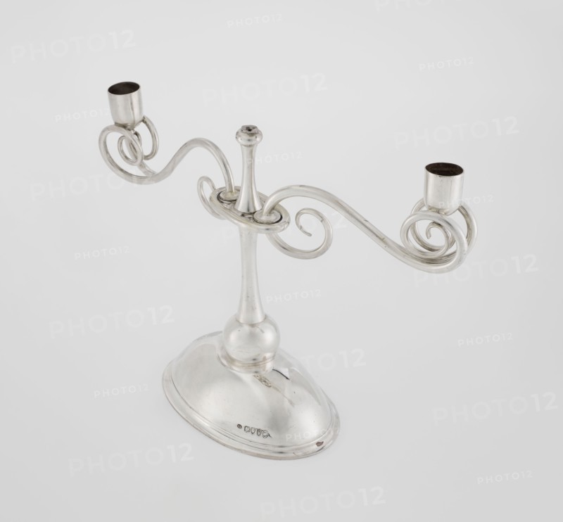 Two-candle candlestick by William Leuchars fl. 1876–1888 and Leuchars & Son London; goldsmith's firm, fl. ca. 1845–1902. Two-branched candlestick on a circular, domed foot. The shaft rests on a ball no finial, enclosed at three-quarters of its height in a plate-like ring, from which extend movable arms made of S-shaped twisted wires, split at the ends, terminating in cylindrical sockets for candles. The foot is engraved with the Pilawa coat of arms with the motto beneath a nine-baton count's crown. Materials/technique: scrap metal. Date: 1881/1882. Place of origin: London, England, Great Britain. Function: lighting. Dimensions: height 27 cm; width 32 cm; depth 11.5 cm; weight 1082 g., A silver-colored metal candelabrum with a rounded base and a tall, tapered central stem supporting a pair of ornate, curving arms that end in two cup-shaped holders; the arms form decorative spirals and loops and the metal surface appears reflective with some darker interior areas in the holders, set agai...