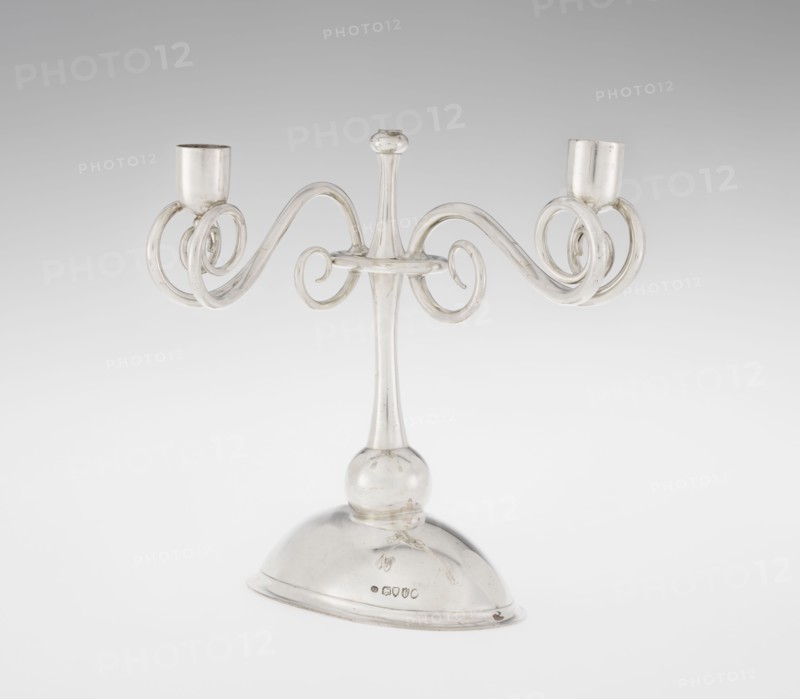 Two-candle candlestick, William Leuchars fl. 1876–1888, Leuchars & Son London; goldsmith's firm, fl. ca. 1845–1902. Two-branched candlestick on a circular domed foot. The shaft rests on a ball no finial, enclosed at three-quarters of its height by a plate-like ring from which extend movable arms made of S-shaped twisted wires, split at the ends, terminating in cylindrical candle sockets. The foot is engraved with the Pilawa coat of arms with the motto beneath a nine-baton count's crown. 1881–1882. England, London. Lighting object. Height 27 cm; width 32 cm; depth 11.5 cm; weight 1082 g., A clear glass candelabrum with a central stem rising from a rounded glass base, featuring a bulbous joint near the base and a slender neck topped by a small rounded finial, with two symmetrically curved arms that form ornate spiral loops ending in cylindrical candle cups; the colors visible are mainly transparent glass with reflections showing shades of white, pale gray, and subtle metallic highlights.
