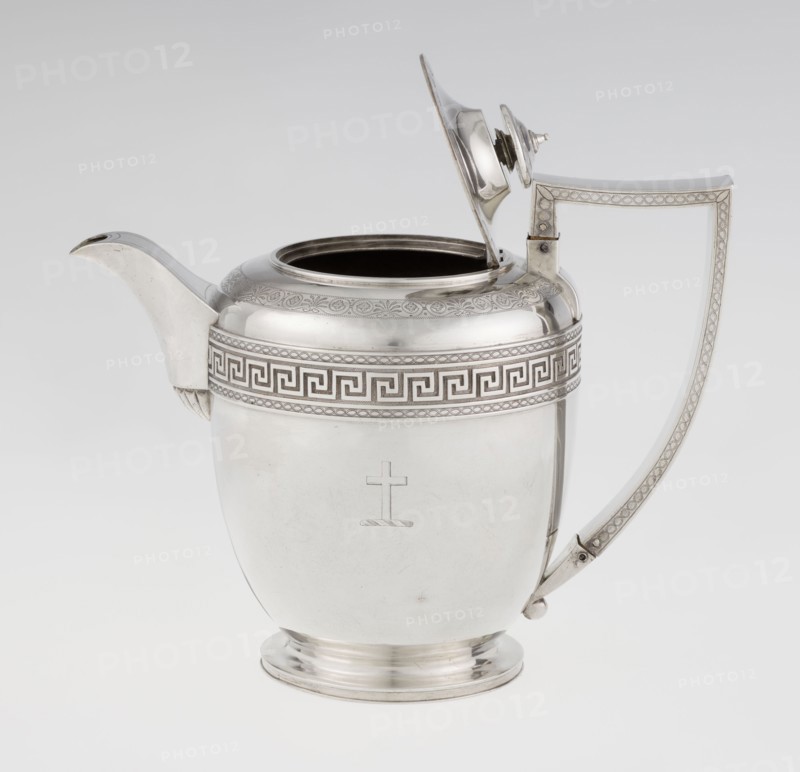 Teapot by John Emes fl. 1796–1808, goldsmith. Ovoid teapot on a circular base with lid, decorated on the upper belly with a wide band of meander ornament and braid. Side features a ribbon-shaped, broken handle decorated with braid; high-set spout decorated at the base with a stylised palmette; lid finished with a knob-shaped handle. Belly engraved with a coat of arms on one side and a cross on the other. Empire/Georgian/George III style, circa 1806–1807, London, England. Medium: silver vessel. Height 17.9 cm; width 23.7 cm; weight 815 g., A polished metallic jug with a reflective silver surface sits against a pale background, featuring a curved spout on one side and an angular handle on the other; the lid is open and attached by a hinge, showing a dark interior rim. The body of the jug has a decorative band near the top with a repeating geometric pattern and a subtle engraved border, and the lower body is smooth with a small simple engraved emblem. The handle and spout have matching de...