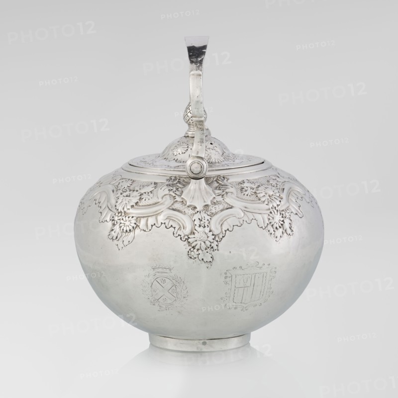 Buliera kettle for boiling water by Benjamin Gignac fl. 1745–1774, goldsmith. Rococo/rocaille style with Georgian/George II influences. Spherical buliera with a flat bow mounted on volutes; spout-shaped side spout; centrally arched cover topped with a pine cone. Decorated with repoussé rococo ornament and floral/plant motifs. Two engraved coats of arms among the decoration — a four-field one in an oval beneath a baronial crown and a second illegible shield. Missing base with a heater. Dated 1753/1754, London, England, Great Britain. Vessel; height 22 cm, width 24 cm, depth 19 cm; weight 848 g., A rounded metal vessel with a hinged domed lid and a curved handle extending from the top is shown against a plain light background. The vessel has ornate raised decorative motifs around the upper portion, including flowing leaf and scroll shapes with textured detailing. The lower surface of the vessel is smooth and reflective with subtle variations in sheen. The handle and lid have matching dec...