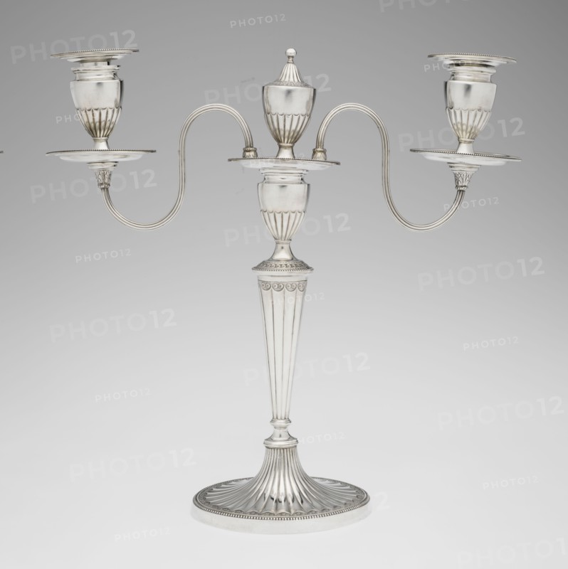 Two-candle candelabrum by Charles Hougham fl. 1769–1793. Two-armed candelabrum on an oval foot, bordered with pearling and decorated with a knobbed rosette. Conical, fluted stem topped with a vase-shaped socket, with a setting of two S-shaped arms terminating in saucers and vase-shaped sockets; in the centre between the arms is a vase topped with a ball. Sleeves decorated halfway with vertical flutes, smooth above. Classicism/George III/Georgian style; fluted, pearling, rosette ornament. Dated 1787/1788. England, London. Lighting. Height 38 cm; width 39 cm; depth 12 cm; weight 1060 g., A silver-colored three-armed candelabrum with a fluted round base and a tall, tapered fluted stem leading to a central urn-shaped holder topped by a removable lid, the central platform connecting to two curved, symmetrical arms that extend outward to hold a group of three cup-shaped candle sockets each set above a round drip plate; the metal surface is smooth and reflective with decorative fluting and be...