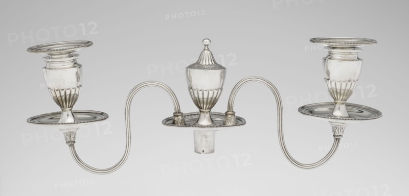 Two-candle candelabrum by Charles Hougham fl. 1769–1793, goldsmith, classicism/George III/Georgian style, 1787–1788, London, England. Two-armed candelabrum on an oval foot bordered with pearling and decorated with a knobbed rosette. Conical, fluted stem topped with a vase-shaped socket that holds a setting with two S-shaped arms terminating in saucers and vase-shaped sockets; central vase topped with a ball. Sleeves decorated halfway with vertical flutes, smooth above. Materials: metal silver; function: lighting. Dimensions: height 38 cm, width 39 cm, depth 12 cm; weight 1060 g., A silver metallic three-armed candleholder with a central covered cup and two matching side cups connected by curved arms, all with rounded bases and fluted lower sections; the surface is reflective with highlights and subtle shadows, showing tones of silver, gray, and off-white, set against a plain light gray background.