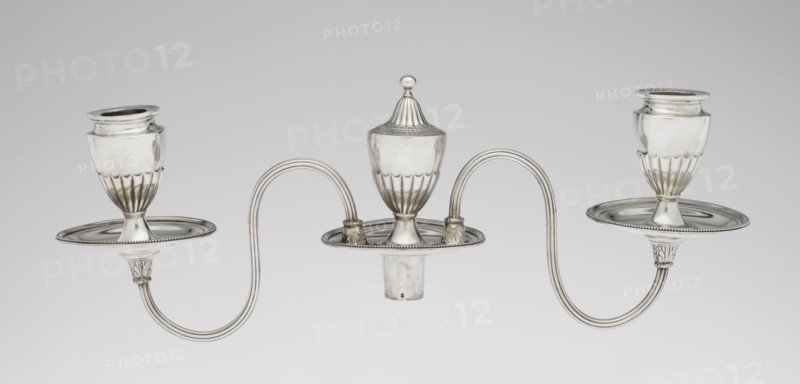 Two-candle candelabrum, Hougham, Charles fl. 1769–1793 goldsmith, classicism, George III / Georgian style. Two-armed candelabrum on an oval foot bordered with pearling and decorated with a knobbed rosette. Conical, fluted stem topped with a vase-shaped socket, containing a setting with two S-shaped arms terminating in saucers and vase-shaped sockets; in the centre between the arms is a vase topped with a ball. Sleeves decorated halfway with vertical flutes, smooth above. Scrap - purchased. 1787/1788. England, London. Function: lighting. Height 38 cm; width 39 cm; depth 12 cm; weight 1060 g., A silver three-armed ornamental object with an overall metallic sheen consisting of a central urn-shaped piece topped by a domed finial and two matching goblet-like holders on either side, connected by gracefully curved tubular arms that form symmetrical loops; each holder sits on a shallow round drip plate with beaded edges, and the bases of the supports feature fluted and acanthus-like decorative...