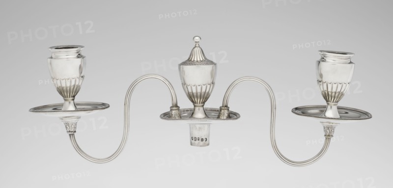 Two-candle candelabrum. Maker: Charles Hougham fl. 1769–1793. Style: Classicism, George III / Georgian. Description: Two-armed candelabrum on an oval foot bordered with pearling and decorated with a knobbed rosette. Conical, fluted stem topped with a vase-shaped socket, containing a setting with two S-shaped arms terminating in saucers and vase-shaped sockets with drip-pans; center between the arms is a vase topped with a ball. Sleeves decorated halfway with vertical flutes, smooth above. Date: 1787/1788. Place: London, England. Function: Lighting. Materials/technique: goldsmith work. Dimensions: height 38 cm, width 39 cm, depth 12 cm; weight 1060 g., A symmetrical metallic object with a shiny silver finish consisting of a central lidded vessel with a rounded finial on top flanked by two matching goblet-like cups, all set on shallow oval trays with beaded rims; the cups and central vessel have fluted lower sections and sit on short stems with decorated collars, and two sinuous double-a...