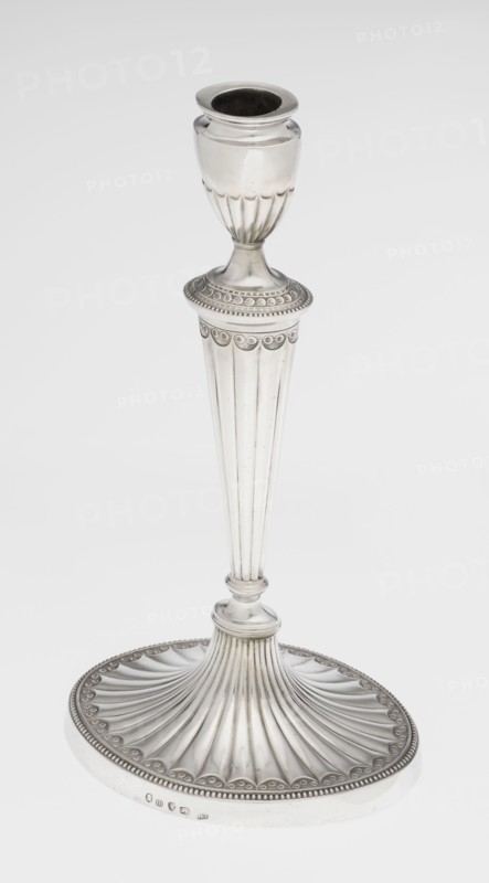 Two-candle candelabrum, maker: Charles Hougham fl. 1769–1793. Classicism/Georgian, George III style. Silver; fluted, pearling and rosette ornament. Two-armed candelabrum on an oval foot bordered with pearling and decorated with a knobbed rosette. Conical fluted stem topped with a vase-shaped socket containing a setting with two S-shaped arms terminating in saucers and vase-shaped sockets; central vase topped with a ball; sleeves decorated halfway with vertical flutes, smooth above. Dated 1787/1788. England, London. Function: lighting. Height 38 cm; width 39 cm; depth 12 cm; weight 1060 g., A tall metallic candlestick with a reflective silver surface standing on an ornate round base; the base has a fluted, fan-like pattern radiating outward and a beaded rim with a repeating scalloped motif, transitioning into a slender, vertically grooved stem that tapers upward to a decorative collar with circular embossed details, leading to a fluted cup-shaped holder at the top. The visible colors ar...