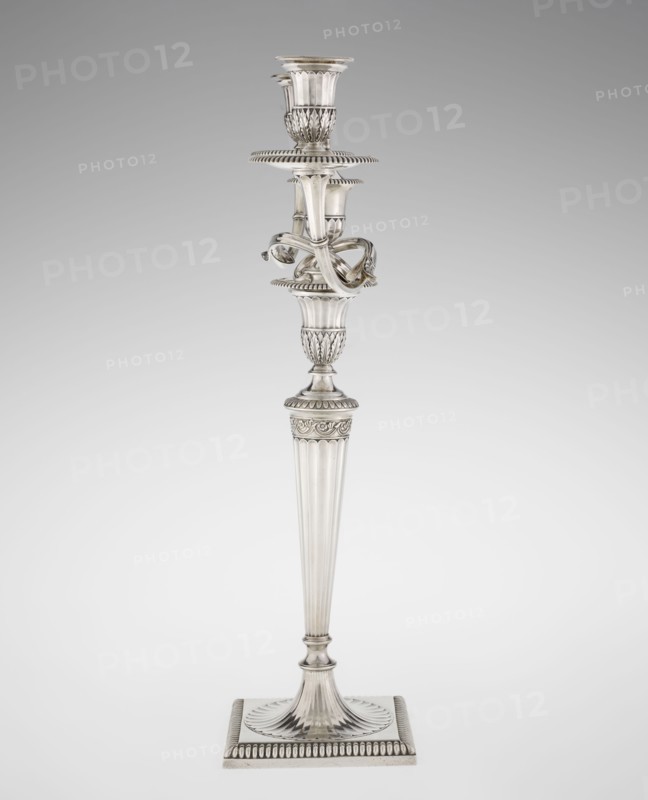 Classicist Two-Armed Candelabra, Schofield, John fl. 1776–1796 goldsmith, classicism style, godron ornaments, monograms, faceting, leaves ornament, Georgian, George III, Classicist two-armed candelabra. On a square base surrounded by godron ornament, passing into a circular foot decorated with a surrounding rosette; faceted stem enclosed at the top with a knurled floral tendril, terminating in a vase-shaped socket decorated with acanthus leaves and facets, edged with godrons, in which is set a setting in the form of two faceted, rotating arms ending with plate-shaped profiled dishes and vase-shaped candle holders, separated by a decorative vase with a lid. Engraved on several elements with a tied monogram PHB; dated 1786/1787, London, England, Great Britain. Function: lighting. Dimensions: height 47.5 cm, width 38 cm, depth 13 cm; weight 850 g., A tall metallic candlestick stands against a plain light gray background, showing a reflective silver surface with polished highlights and sub...