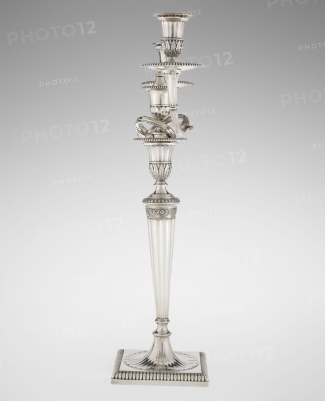 Classicist Two-Armed Candelabra, Schofield, John fl. 1776–1796 goldsmith; classicism, Georgian/George III style. Classicist two-armed candelabra on a square base surrounded by godron ornament, passing into a circular foot decorated with a surrounding rosette; faceted stem enclosed at the top with a knurled floral tendril, terminating in a vase-shaped socket decorated with acanthus leaves and facets, edged with godrons, in which is set a setting in the form of two faceted, rotating arms ending with plate-shaped profils and vase-shaped candle holders, separated by a decorative vase with a lid. Engraved on several elements with a tied monogram PHB, dated 1786/1787, London, England, Great Britain. Function: lighting. Dimensions: height 47.5 cm, width 38 cm, depth 13 cm; weight 850 g., A tall ornate metal candlestick stands against a neutral gray background; it has a square base with fluted and beaded edges, a tapered fluted column rising to an intricately molded central section with acanth...