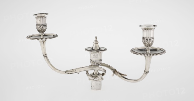 Classicist Two-Armed Candelabra, Schofield, John fl. 1776–1796 goldsmith, classicism, godron ornaments, monograms, faceting, leaves ornament, Georgian, George III. Classicist two-armed candelabra on a square base surrounded by godron ornament, passing into a circular foot decorated with a surrounding rosette; faceted stem enclosed at the top with a knurled floral tendril, terminating in a vase-shaped socket decorated with acanthus leaves and facets, edged with godrons, in which is set a setting in the form of two faceted, rotating arms ending with plate-shaped profils and vase-shaped candle holders, separated by a decorative vase with a lid. Engraved on several elements with a tied monogram PHB, 1786/1787, London, England, Great Britain. Function: lighting. Height 47.5 cm, width 38 cm, depth 13 cm, weight 850 g., A metallic three-arm candle holder with a central finial and two outward-curving arms ending in raised circular drip plates and cup-shaped holders; the metal surface is polish...