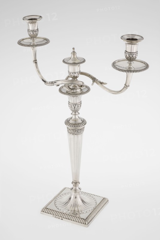 Classicist Two-Armed Candelabra, Schofield, John fl. 1776–1796 goldsmith; classicism, Georgian/George III styles. Classicist two-armed candelabra on a square base surrounded by godron ornament, passing into a circular foot decorated with a surrounding rosette; faceted stem enclosed at the top with a knurled floral tendril, terminating in a vase-shaped socket decorated with acanthus leaves and facets, edged with godrons, in which is set a setting in the form of two faceted, rotating arms ending with plate-shaped profilers and vase-shaped candle holders, separated by a decorative vase with a lid. Engraved on several elements with a tied monogram PHB, dated 1786/1787, London, England. Lighting; height 47.5 cm, width 38 cm, depth 13 cm, weight 850 g., A silver-colored three-armed candelabrum stands on a square base with ridged edging and a fluted, scalloped central platform; a tall, tapered fluted column rises from the base, topped by an ornate capital with ribbed and leaf-like patterns, s...
