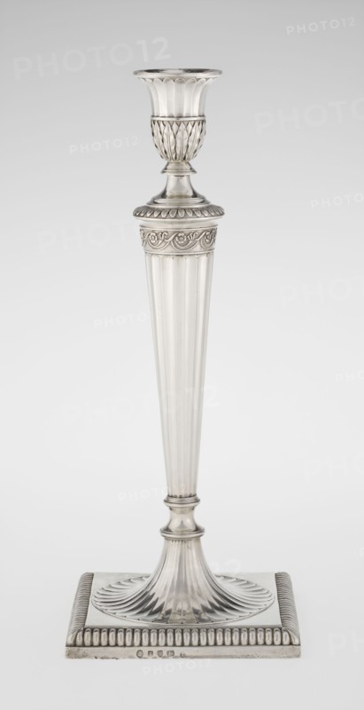 Classicist Two-Armed Candelabra, Schofield, John fl. 1776–1796 goldsmith, classicism, Georgian / George III style. Classicist two-armed candelabra on a square base with godron ornament, passing into a circular foot decorated with a surrounding rosette; faceted stem enclosed at the top with a knurled floral tendril, terminating in a vase-shaped socket decorated with acanthus leaves and facets, edged with godrons. Set in the socket are two faceted, rotating arms ending with plate-shaped profits and vase-shaped candle holders, separated by a decorative vase with a lid. Engraved on several elements with a tied monogram PHB; dated 1786/1787. Place: London, England. Function: lighting. Dimensions: height 47.5 cm, width 38 cm, depth 13 cm; weight 850 g., A single tall metallic candlestick stands against a plain light background, rendered in silver tones with reflective highlights and subtle darker gray shadows. The candlestick has a flared cup at the top with vertical fluting, a decorative te...