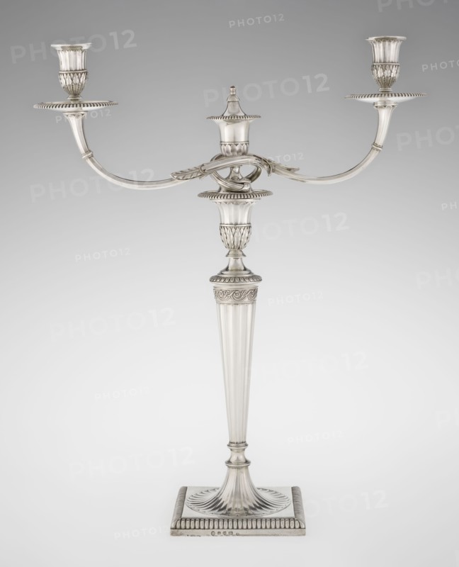 Classicist Two-Armed Candelabra, by John Schofield fl. 1776–1796, goldsmith; Classicism/Georgian/George III style. Classicist two-armed candelabra on a square base surrounded by godron ornament, passing into a circular foot decorated with a surrounding rosette; faceted stem enclosed at the top with a knurled floral tendril, terminating in a vase-shaped socket decorated with acanthus leaves and facets, edged with godrons, in which is set a setting in the form of two faceted, rotating arms ending with plate-shaped profils and vase-shaped candle holders, separated by a decorative vase with a lid. Decorated with godron ornaments, faceting, leaves ornament and engraved on several elements with a tied monogram PHB, dated 1786/1787, London, Great Britain. Function: lighting. Dimensions: height 47.5 cm, width 38 cm, depth 13 cm; weight 850 g., A silver candelabrum stands against a neutral light gray background, featuring a tall fluted central stem rising from a square base with ridged edges an...