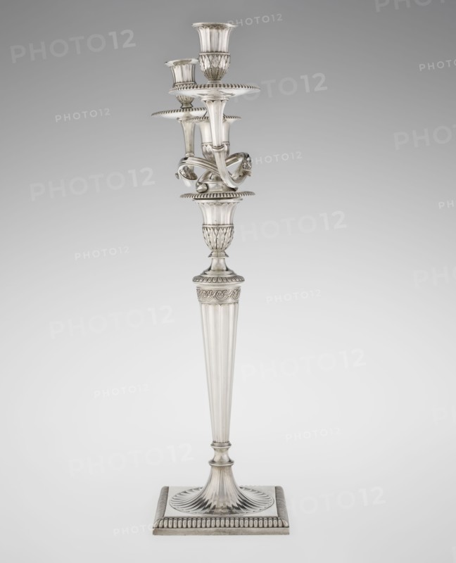 Classicist Two-Armed Candelabra, Schofield, John fl. 1776–1796 goldsmith, classicism style, godron ornaments, monograms, faceting, leaves ornament, Georgian, George III. Classicist two-armed candelabra on a square base surrounded by godron ornament, passing into a circular foot decorated with a surrounding rosette; faceted stem enclosed at the top with a knurled floral tendril, terminating in a vase-shaped socket decorated with acanthus leaves and facets, edged with godrons, in which is set a setting in the form of two faceted, rotating arms ending with plate-shaped profits and vase-shaped candle holders, separated by a decorative vase with a lid. Engraved on several elements with a tied monogram PHB, 1786/1787, England, London, Great Britain; function: lighting. Height 47.5 cm, width 38 cm, depth 13 cm, weight 850 g., A tall ornate metal candlestick stands against a plain background, with a fluted tapered shaft rising from a square base that has a ridged border and a circular fluted p...