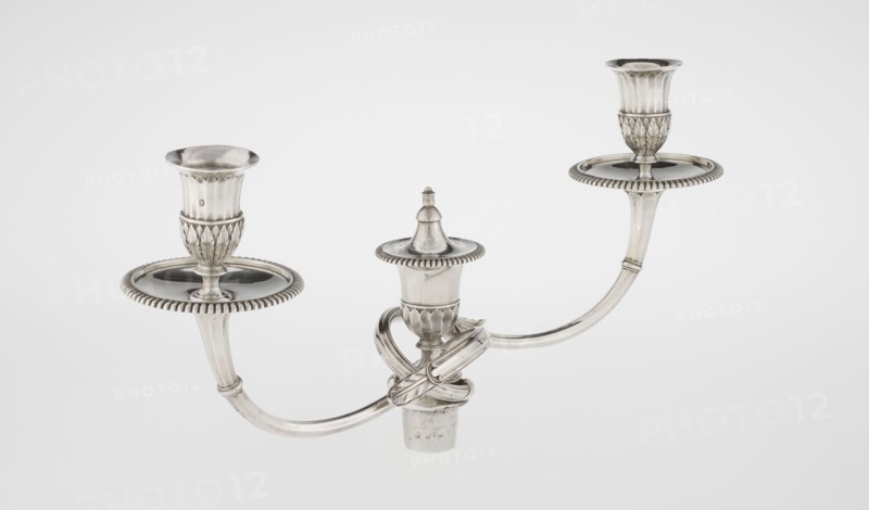 Classicist Two-Armed Candelabra, Schofield, John fl. 1776–1796 goldsmith; classicism, Georgian/George III style. Classicist two-armed candelabra on a square base surrounded by godron ornament, passing into a circular foot decorated with a surrounding rosette; faceted stem enclosed at the top with a knurled floral tendril, terminating in a vase-shaped socket decorated with acanthus leaves and facets, edged with godrons, in which is set a setting in the form of two faceted, rotating arms ending with plate-shaped profils and vase-shaped candle holders, separated by a decorative vase with a lid. Engraved on several elements with a tied monogram PHB, dated 1786/1787, London, England. Function: lighting. Dimensions: height 47.5 cm, width 38 cm, depth 13 cm; weight 850 g., A metallic three-armed candelabrum with a polished silver surface, showing a central decorative finial and two matching candle cups on curved arms, each cup sitting on a ridged circular drip tray; the design includes fluted...