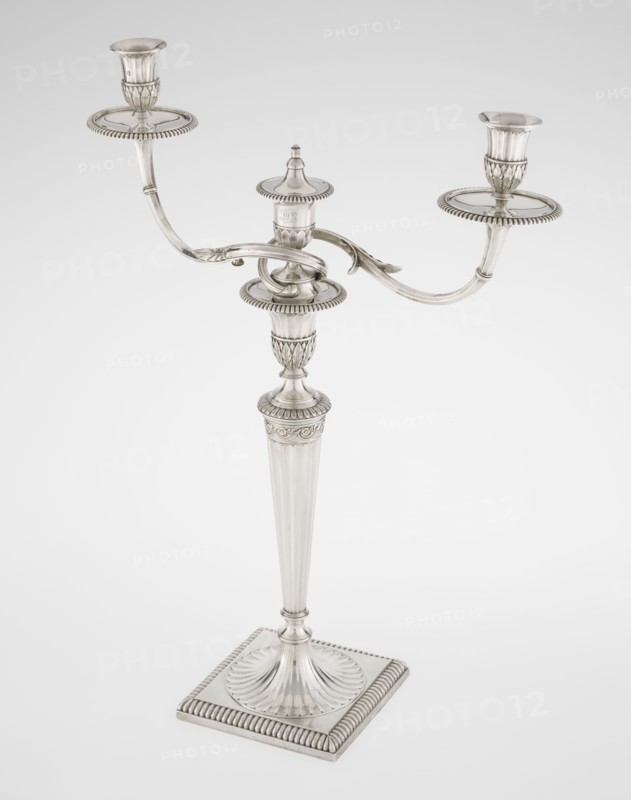 Classicist two-armed candelabra by John Schofield fl. 1776–1796, goldsmith. Classicism/Georgian George III style. On a square base surrounded by godron ornament, passing into a circular foot decorated with a surrounding rosette; faceted stem enclosed at the top with a knurled floral tendril, terminating in a vase-shaped socket decorated with acanthus leaves and facets, edged with godrons, containing two faceted, rotating arms ending with plate-shaped profiled and vase-shaped candle holders, separated by a decorative vase with a lid. Engraved on several elements with a tied monogram PHB, dated 1786/1787, London, England. Material: silver lighting. Dimensions: height 47.5 cm, width 38 cm, depth 13 cm; weight 850 g., A silver-colored three-armed candelabrum standing on a square base with fluted and beaded decorative edges, a tapered fluted central column rising to an ornate central socket, and two curved arms extending outward to hold cup-shaped candle sockets atop round drip trays, all s...