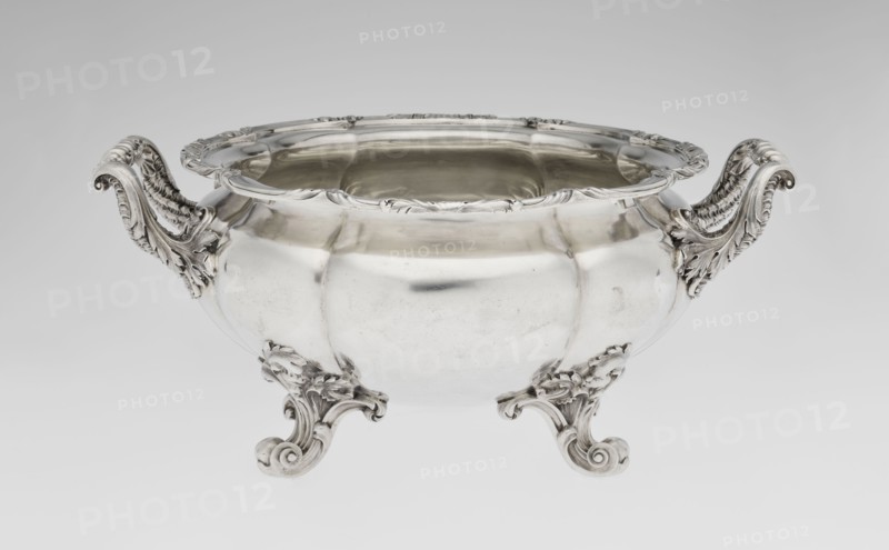 Punch vase by Paul Storr 1771–1844 and Storr & Mortimer London; goldsmiths, fl. 1822–1838. English, 1831/1832. Silver punch vessel, spherical flattened eight-part, burled and ribbed with burling and ribbing; supported on four volute leafy feet. Two handles decorated with acanthus leaves and stylized shells. Upper edge turned up and decorated with relief ornament of leaves and shells. Belly engraved with a coat of arms in a decorative cartouche. Materials/technique: silver; motifs include acanthus, shells, volutes. Dimensions: height 15.5 cm, width 36.2 cm, depth 27.8 cm; weight 2490 g., A shiny silver bowl with ornate scrolling handles and feet sits against a pale gray background, the bowl's rounded body reflecting light and showing smooth and slightly dimpled surfaces, the rim decorated with carved floral and leaf motifs, the handles and feet featuring elaborate acanthus-like scrollwork and textured detailing in the same silver tone, and the overall colors visible are silver and shade...