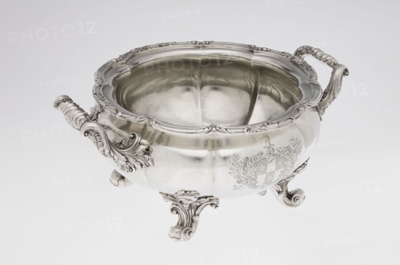 Punch vase, maker: Paul Storr 1771–1844; firm: Storr & Mortimer London; goldsmiths, fl. 1822–1838. Spherical, flattened, eight-part burled and ribbed punch vase supported on four volute leafy feet. Two handles decorated with acanthus leaves and stylized shells. Upper edge turned up and decorated with a relief ornament of leaves and shells. Belly with an engraved coat of arms in a decorative cartouche. Materials/technique: burling. Subjects/ornament: acanthus, shells, volutes, coats of arms. Date: 1831/1832. Place of origin: London, England, Great Britain. Function: punch vessel. Dimensions: height 15.5 cm; width 36.2 cm; depth 27.8 cm; weight 2490 g., A decorative metal bowl with an ornate rim and curved sides sits on a smooth light background, showing a reflective silver surface with polished highlights and soft shadows; it has a pair of elaborately carved handles with scroll and leaf motifs and rests on ornate curled feet with similar decorative detailing, and the interior reflects t...