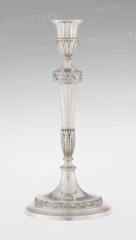 Single-candle candlestick by John Schofield fl. 1776–1796. Classicism/Georgian/George III style. Silver candlestick supported on a circular, stepped-profile foot, decorated with a band of stylised acanthus tendrils with flowers; faceted stem, enclosed at the bottom by lanceolate leaves, widening towards the top and terminating in a ring knurled with oak leaves; vase sleeve with lanceolate leaves at the bottom, faceted above with a slightly rolled-up edge. Dated 1789/1790. England, London. Function: lighting. Height 29.8 cm; diameter 13.8 cm; weight approx. 680 g., A single tall metallic object with a reflective silver surface stands against a light gray background, featuring a flared top with vertical ridges, a rounded midsection with carved leaf-like patterns, a long tapered column with fluted vertical grooves, a narrower ribbed section with elongated leaf motifs, and a broad circular base decorated with a raised band of floral or vine ornamentation; the visible colors are silver and ...