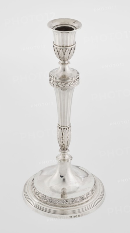 Single-candle candlestick by John Schofield fl. 1776–1796. Classicism/Georgian/George III style. Silver candlestick supported on a circular stepped-profile foot, decorated with a band of stylised acanthus tendrils with flowers; faceted stem enclosed at the bottom by lanceolate leaves, widening toward the top and terminating in a ring knurled with oak leaves; vase sleeve with lanceolate leaves at the bottom, faceted above with a slightly rolled-up edge. Inscribed/dated 1789/1790. England, London. Function: lighting. Height 29.8 cm; diameter 13.8 cm; weight approx. 680 g., A single tall metallic candlestick with a polished silver surface, showing ornamental fluted and leaf-like carved sections along its stem and base; the top has a flared cup with a dark interior, the middle features a rounded knop with a decorative band, and the wide circular base has a raised domed center and a patterned border. The visible colors are silver, metallic gray, and dark gray inside the cup.