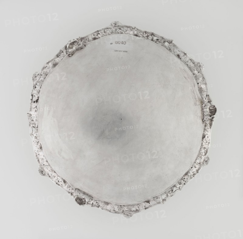 Leg tray by Robert Harper fl. 1853–1869, maker; Harper & Son London, goldsmith's firm, fl. 1869–1889. Circular patera supported on three volute legs terminating in vine leaves at the base. The mirror features a blind reserve richly decorated with engraved Neo-Rococo ornament in the form of rocailles, rooster combs, acanthus volutes, and floral sprigs. The slightly raised, openwork collar features relief decoration of vine leaves and grapes, with four Dionysus masks at the feet, and high-relief insects butterflies, moths, midges, snails, and a caterpillar. The collar edge is profiled with a half-cylinder decorated with shells and a rooster comb. Subject/iconography: head of Dionysus; motifs: insects, vines, snails, caterpillars, rocaille, shells, volutes, acanthus leaf. Date: 1860–1861. Place: London, England, Great Britain. Medium/object type: vessel leg tray. Dimensions: height 3.9 cm; diameter 32.5 cm; weight 895 g., A round metallic tray viewed from above with a smooth central surfa...