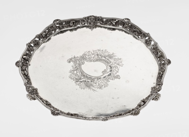 Leg Tray by Robert Harper fl. 1853–1869; Harper & Son London; goldsmith's firm; fl. 1869–1889. Circular patera supported on three volute legs terminating in vine leaves at the base. The mirror features a blind reserve richly decorated with engraved Neo-Rococo ornament in the form of rocailles, rooster combs, acanthus volutes, and floral sprigs. The slightly raised, openwork collar bears relief decoration of vine leaves and grapes, with four Dionysus masks at the feet, and high-relief insects butterflies, moths, midges, snails, and a caterpillar. The collar edge is profiled with a half-cylinder decorated with shells and a rooster comb. Iconography: head of Dionysus. Materials/ornament: insects, vines, snails, caterpillars, rocaille, shells, volutes, acanthus leaf; style: Neo-Rococo. Date: 1860/1861. Place: London, England, Great Britain. Vessel. Height: 3.9 cm; diameter: 32.5 cm; weight: 895 g., A round decorative tray with an ornate raised rim featuring floral and scrollwork motifs and...