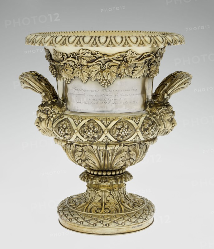 Wine Cooler, Boulton & Plate Co Birmingham; goldsmith and plater firm; fl. 1768–1834 maker; vine ornament; dedications; jockey on horse iconographic; horse racing; classicism style; Georgian style; acanthus leaf ornament; Satyr heads iconographic; cymatium ornament. Classicist on a circular foot decorated with a half-cylinder of laurel and acanthus leaves overlapping a short stem; body enclosed below with large acanthus leaves, above decorated with vines in a diagonal, rhombic braid. Plaque with racers on the front riders; on the back a plaque with a secondary inscription in Cyrillic script is attached the original inscription, probably referring to a prize in horse races, has been erased. Under the upper edge a decorative strip of vines; the rim is edged with a stylized kimation; on the sides massive ears mounted in satyr masks. Date: 1826/1827. Place of manufacture: Birmingham, England, Great Britain. Object type: vessel wine cooler. Dimensions: height 30 cm; width 27 cm; depth 24.5 ...
