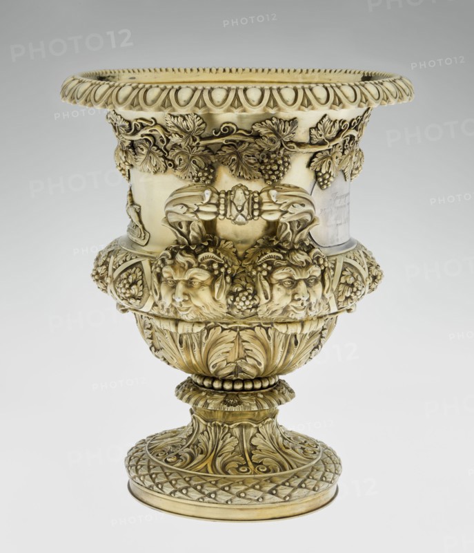 Wine cooler by Boulton & Plate Co. Birmingham; goldsmith and plater firm; fl. 1768–1834. Silver vessel, 1826/1827. Classicism, Georgian style. Circular foot decorated with a half-cylinder of laurel and acanthus leaves overlapping a short stem; body enclosed below with large acanthus leaves, above decorated with diagonal vine in a rhombic braid. Plaque on the front with racers jockey on horse; horse racing; a secondary plaque on the back bears an inscription in Cyrillic script the original inscription, probably referring to a prize in horse races, has been erased. Decorative strip of vines under the upper edge; rim edged with a stylized cymatium/kimation; massive handles mounted in satyr masks. Decorations include vine ornament, acanthus leaf, satyr heads, and classicist motifs. Height 30 cm; width 27 cm; depth 24.5 cm; weight 3725 g., A decorative ornate urn with a metallic golden color overall, featuring a wide flared rim with a repeating scalloped edge and carved teardrop motifs, a b...