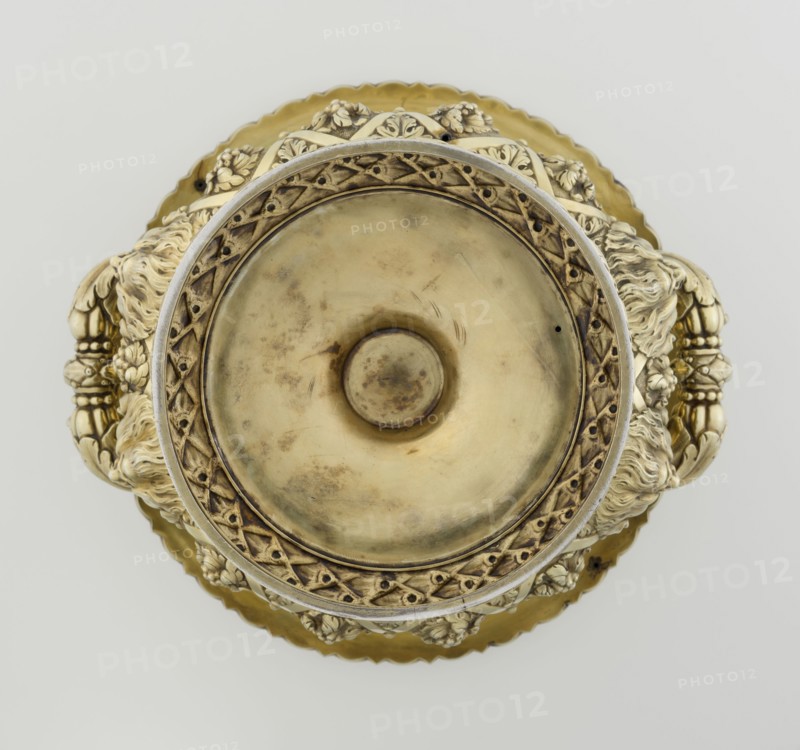 Wine cooler by Boulton & Plate Co Birmingham; goldsmith and plater firm; fl. 1768–1834, 1826/1827. Silver vessel, height 30 cm, width 27 cm, depth 24.5 cm, weight 3725 g. Classicism/Georgian style. Circular foot decorated with a half-cylinder of laurel and overlapping acanthus leaves on a short stem; body enclosed below with large acanthus leaves, above decorated with vines in a diagonal rhombic braid. Plaque with horse racers on the front; a secondary plaque with an inscription in Cyrillic on the back original inscription, probably referring to a prize in horse races, has been erased. Upper edge with a decorative strip of vines; rim edged with a stylized cymatium; massive handles mounted in satyr masks. Ornament: vine, acanthus leaf, cymatium; iconography: jockey on horse, horse racing, satyr heads., A top-down view of an ornate metallic vessel with a central circular depression, surrounded by an intricate raised rim with lattice-like patterns and floral motifs; the piece shows a mix ...