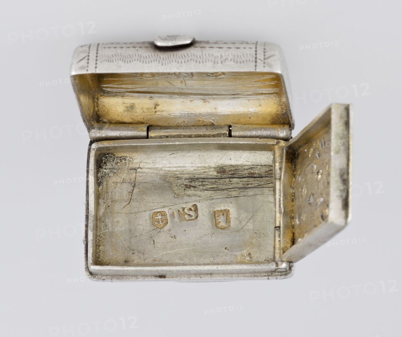 Fragrance container vinaigrette. Maker: Thomas Shaw fl. 1822–ca. 1840; 1782–1845, goldsmith. Scrap - purchase, purchase provenance. Vinaigrette in the form of a bag with an envelope closure and a protruding tongue, decorated on the front with dot-engraved flowers and leaves. Hinged lid opens to reveal an openwork insert decorated with plant scrolls. Materials/technique: engraving, openwork. Date: 1834/1835. Place: Birmingham, England, Great Britain. Function: toilet accessory. Dimensions: height 2.1 cm, width 1.5 cm, depth 1 cm; weight 10 g., A small rectangular metal box with its lid open, showing an inner surface with worn and scratched areas; the interior has a mix of silver and warm gold tones with patches of darker tarnished spots, and the exterior edges appear silver with engraved patterns on the lid. The hinge and inner rim show metallic wear, and the inner side of the lid displays horizontal scratch marks and a golden wash; the right side panel of the box is open and reveals a ...