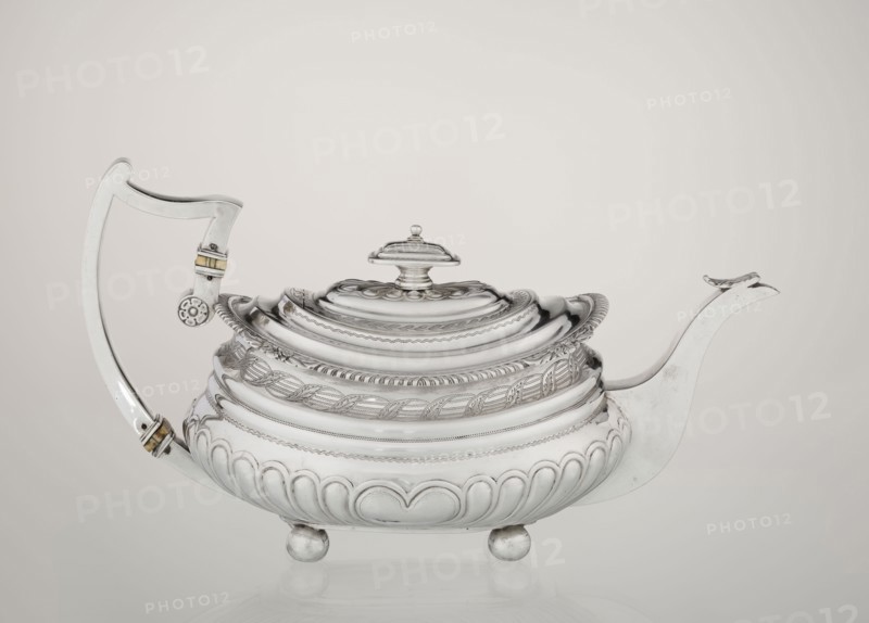 Teapot. Maker: George McHattie fl. 1799–ca. 1825, goldsmith. English Regency style, 1819–1820, Edinburgh, Scotland, Great Britain. Rectangular teapot with rounded edges, supported by four ball feet; decorated with hoardings ornament at the bottom and two smooth half-cylinders above; engraved band of braid at the narrowing; upper edge with a collar decorated with hoardings and flowers. Hinged, rectangular profiled lid edged with tendril; spout-shaped spout set high on the axis; broken ear with bone insulators. Materials/function: tea vessel. Dimensions: height 16 cm, width 28.5 cm, depth 13.5 cm, weight 735 g., A polished silver teapot with an ornate lid and a decorative finial sits on a reflective surface, showing a curved handle on one side and a long spout on the other; the body has layered, rounded ridges and embossed leaf and rope-like patterns, with scalloped and petal motifs near the base and resting on a group of rounded feet; the handle features small bands of a contrasting war...
