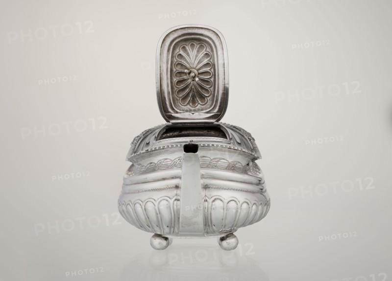 Teapot, maker: George McHattie fl. 1799–ca. 1825. Rectangular teapot with rounded edges, supported by four ball feet. Decorated with hoardings ornament at the bottom, above which are two smooth half-cylinders; at the narrowing an engraved band of braid. Upper edge with a collar decorated with hoardings and flowers. Hinged rectangular profiled lid edged with tendril. Spout-shaped spout set high on the axis; broken ear with bone insulators. English Regency style, 1819–1820. Edinburgh, Scotland, Great Britain. Silver vessel. Height 16 cm, width 28.5 cm, depth 13.5 cm, weight 735 g., A metal vessel with a hinged lid is shown from the front on a neutral background; the lid is open to reveal an embossed pattern on its underside, and the vessel has a rounded, ribbed body raised on a few round feet with a pronounced handle extending toward the viewer; the surface shows decorative engravings and occasional scuffs, and the visible colors are silver, gray, and off-white.