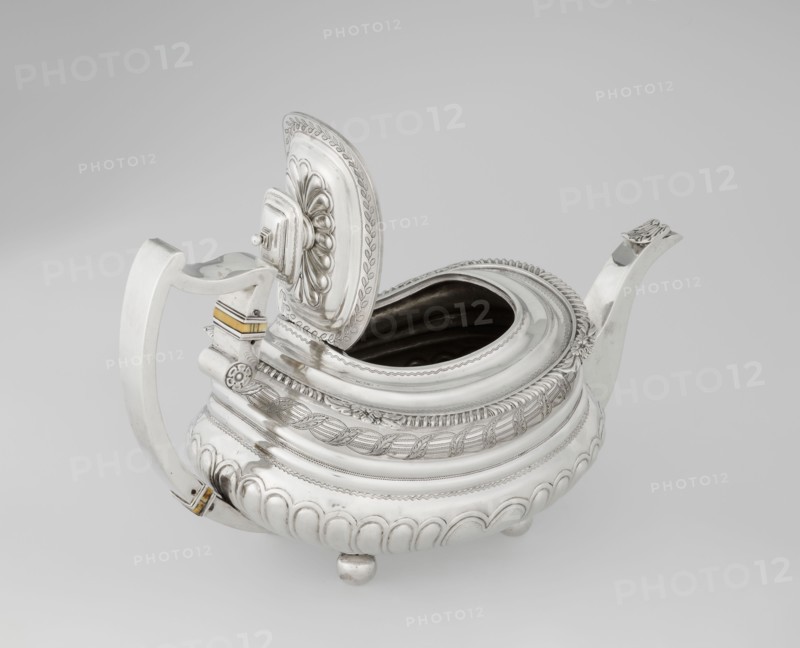 Teapot by George McHattie fl. 1799–ca. 1825, English Regency style. Rectangular teapot with rounded edges, supported by four ball feet. Decorated with hoarding ornament at the base; above are two smooth half-cylinders with an engraved braided band at the narrowing. Upper edge has a collar decorated with hoardings and flowers. Hinged rectangular profiled lid edged with tendril; high axis spout shaped like a spout. Broken ear with bone insulators. Dated 1819–1820, made in Edinburgh, Scotland. Vessel. Height 16 cm; width 28.5 cm; depth 13.5 cm; weight 735 g., A silver teapot with an open hinged lid sits against a neutral background, showing a rounded body with decorative ridges and embossed patterns; the lid has a raised floral-like design and an ornate border, and the interior visible through the opening appears dark. The teapot has a curved spout and a wide handle with small rectangular accents in a yellowish-gold color near its attachments. The body rests on small rounded feet and feat...