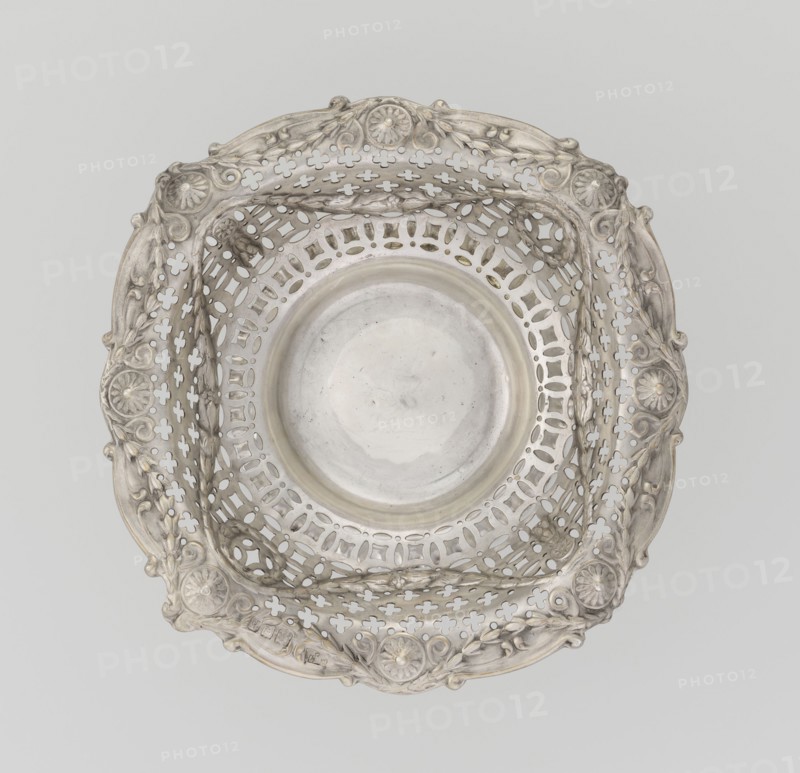 Openwork cake stand by Mappin & Webb Ltd goldsmith, plater, sellers; fl. 1859-. Openwork cake stand on a circular base with a flange, raised and decorated with relief ornament of rocaille and floral motifs; short stem supporting a circular bowl transitioning into a square one with upturned, rounded edges. Edges decorated with ovals, festoons, wreaths and rosettes. Materials/technique: openwork, relief decoration. Date: 1903/1904. Place: London, England. Object type: vessel. Dimensions: height 7.2 cm; width 14.3 cm; depth 14.3 cm; weight 112 g., A decorative metallic bowl with a round smooth center and an ornate pierced square rim, showing a group of floral and scroll motifs around the edges; the surface appears shiny and reflective with areas of subtle tarnish, and the visible colors are silver and light grey.