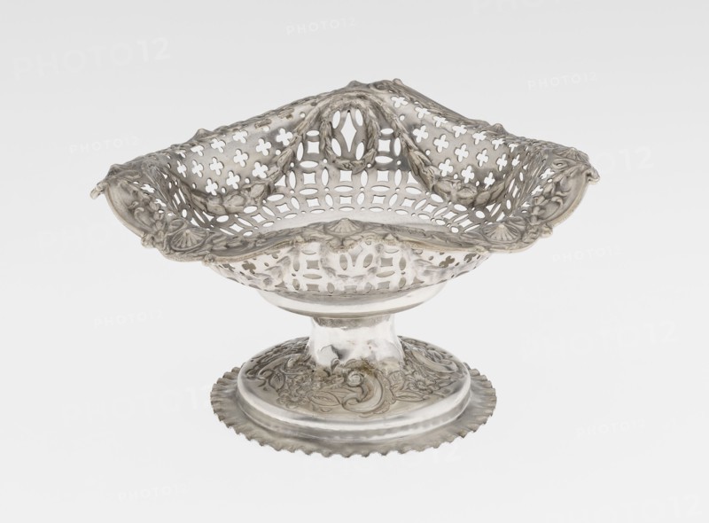 Openwork cake stand by Mappin & Webb Ltd goldsmith, plater, sellers; fl. 1859-. Openwork cake stand on a circular base with a flange, raised and decorated in relief with rocaille and floral motifs. Short stem supports a circular bowl transitioning into a square one with upturned, rounded edges, decorated with ovals, festoons, wreaths and rosettes. Materials/technique: openwork, relief decoration. Date: 1903/1904. Place: London, England. Object type: vessel. Dimensions: height 7.2 cm; width 14.3 cm; depth 14.3 cm; weight 112 g., A decorative metal bowl with an ornate pierced lattice design and scalloped rim sits on a round pedestal base; the bowl and base show detailed embossed floral and scroll motifs and a draped, wavy edge. The object appears in metallic silver tones with areas of darker patina and subtle reflections, set against a plain light gray to white background.
