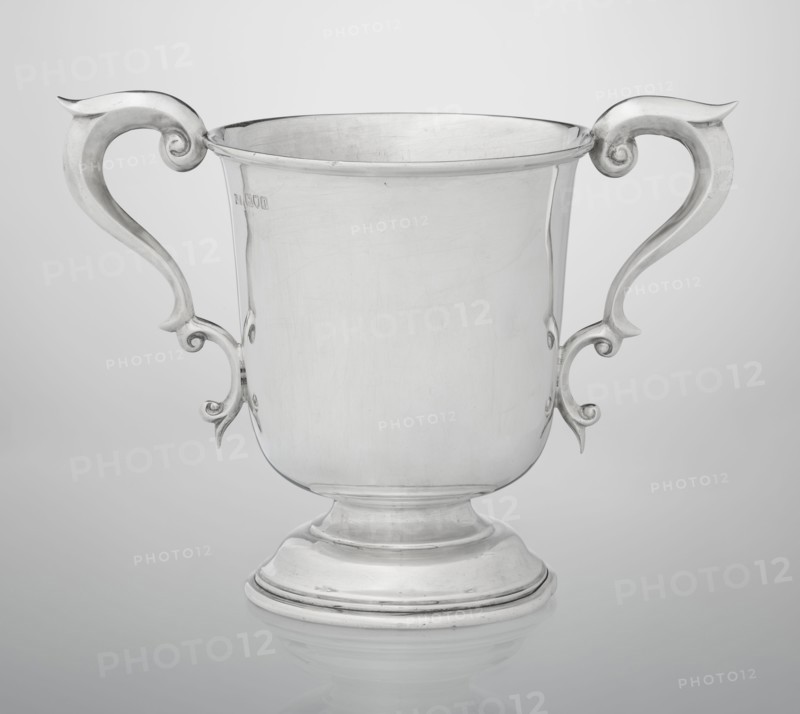 Prize cup by Mappin & Webb Ltd goldsmith, silversmith, dealer; fl. 1859-. Cup on a circular, arched, profiled base with a short shaft; semi-ovoid body flattened at the bottom; slightly flared upper edge bordered with a half-cylinder and two volute handles on the sides. Engraved on the belly: BURSTON POINT TO POINT MEETING / 22nd APRIL 1911 / OPEN STEEP LECHASE / WON BY / MR H.DETERDING'S / 'NIGGER'. Dated 1910/1911. England, London. Vessel; height 16.5 cm, width 23.5 cm, depth 14.5 cm, weight 500 g., A polished silver-colored metal trophy cup with a rounded footed base and a wide cylindrical body, shown front-facing against a light gray background; the cup has a pair of symmetrical ornate handles with curved scroll-like shapes extending outward and upward, and the surface of the metal shows subtle scratches and reflections.