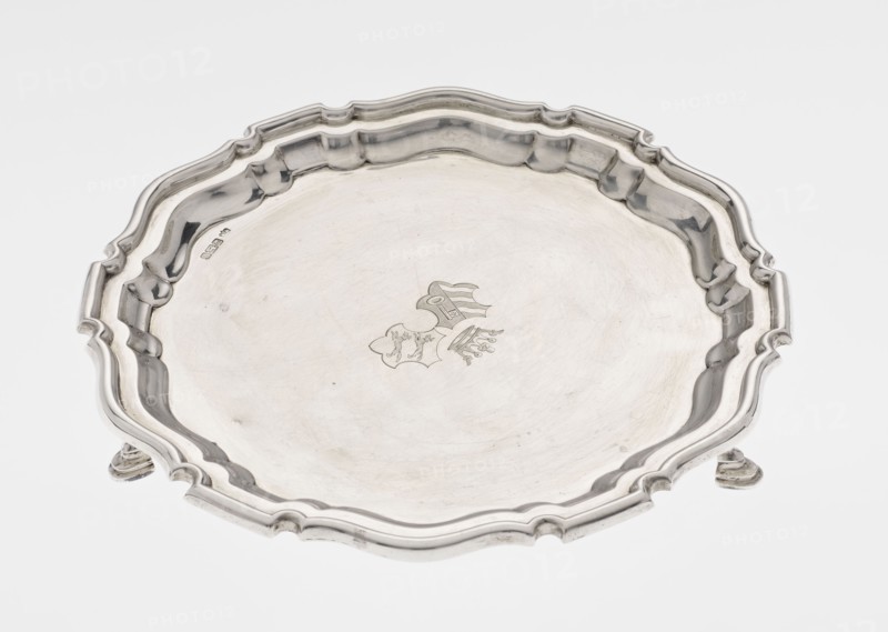 Tray on legs, goldsmith/silversmith maker: Mappin & Webb Ltd. Circular tray supported on three hoof-shaped legs, with a slightly raised, multi-partite profiled and ribbed collar with serrated edges. Ribbed surface; two coats of arms engraved in the mirror beneath a noble crown. Inscription in italics on the bottom center: 29.9.1906 /von HvG. Date: 1906/1907. Material: presumably silver table accessory. Dimensions: height 2.4 cm, diameter 21.2 cm, weight 350 g. Location made: Sheffield, England., A shallow decorative metal tray with a scalloped, ornate rim and a slightly raised center, shown from above; the tray has a smooth reflective surface with subtle scratches and a faint engraved emblem near the center; the rim has molded curves and a few raised sections and stands on small feet that are partly visible; the visible colors are silver tones, gray, and off-white from reflections.