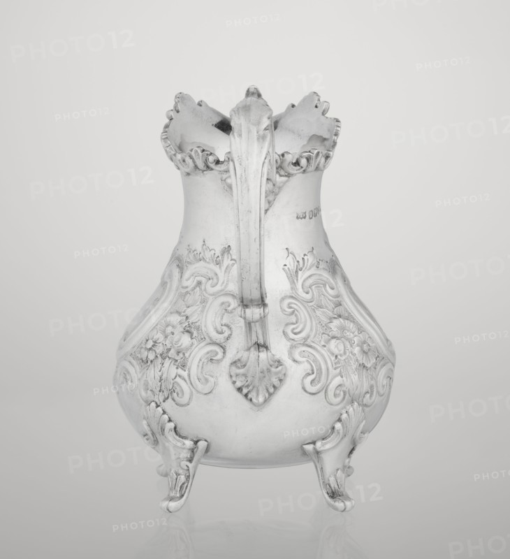 Milk jug by Mappin & Webb Ltd goldsmith/silversmith, neo-rococo style, 1895–1896. Pear-shaped milk jug supported on four volute legs, decorated with an acanthus leaf at the base; belly ornamented with rococo motifs including channel-shaped jars, rocailles, flowers and acanthus leaves. Volute handle with acanthus projections and a wide raised spout with serrated edge. Material: metal. Place: Sheffield, England. Function: vessel. Dimensions: height 14.4 cm, width 9.5 cm, depth 8.5 cm; weight 266 g., A silver-colored ornate pitcher with a scalloped rim and a single curved handle, decorated with raised floral and scrollwork patterns on its bulbous body and standing on a few decorative feet, shown against a light gray background.