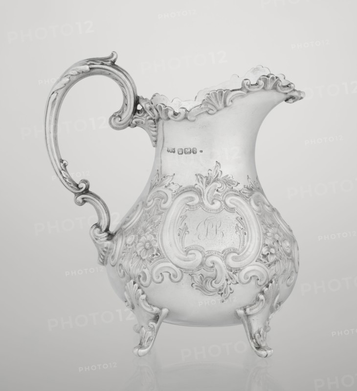 Milk jug by Mappin & Webb Ltd goldsmith, silversmith, dealer; fl. 1859–, neo-rococo style, pear-shaped milk jug supported on four volute legs, decorated with an acanthus leaf at the base and on the belly with rococo motifs in the form of channel-shaped jars, rocailles, flowers and acanthus leaves; volute handle with acanthus projections and a wide, raised spout with a serrated edge; 1895–1896; Sheffield, England, Great Britain; vessel; height 14.4 cm, width 9.5 cm, depth 8.5 cm; weight 266 g., A single ornate metal pitcher with a curved handle and decorative scalloped rim sits against a plain background; the surface shows elaborate raised floral and scrollwork patterns across its rounded body and feet, and the handle features curled, carved details; the image appears in shades of gray, silver, and white.