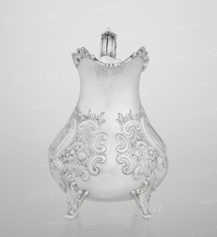 Milk jug, goldsmith/silversmith/dealer Mappin & Webb Ltd fl. 1859– maker, scrap - neo-rococo style, rocaille ornament, flowers, channel-shaped jars ornament, acanthus leaves ornament; pear-shaped milk jug supported on four volute legs, decorated with an acanthus leaf at the base, decorated on the belly with rococo motifs in the form of channel-shaped jars, rocailles and flowers and acanthus leaves; volute handle with acanthus projections and a wide, raised spout with a serrated edge; 1895–1896; Sheffield, England, Great Britain; vessel; height 14.4 cm, width 9.5 cm, depth 8.5 cm, weight 266 g, A decorative pitcher with an ornate, curving silhouette sits against a plain background, displaying a smooth upper body that flares into a scalloped rim and a detailed handle rising from the back; the lower body features embossed floral and scrollwork patterns and rests on three cabriole-style feet with curled toes. The visible colors are shades of gray, silver, white, and off-white.