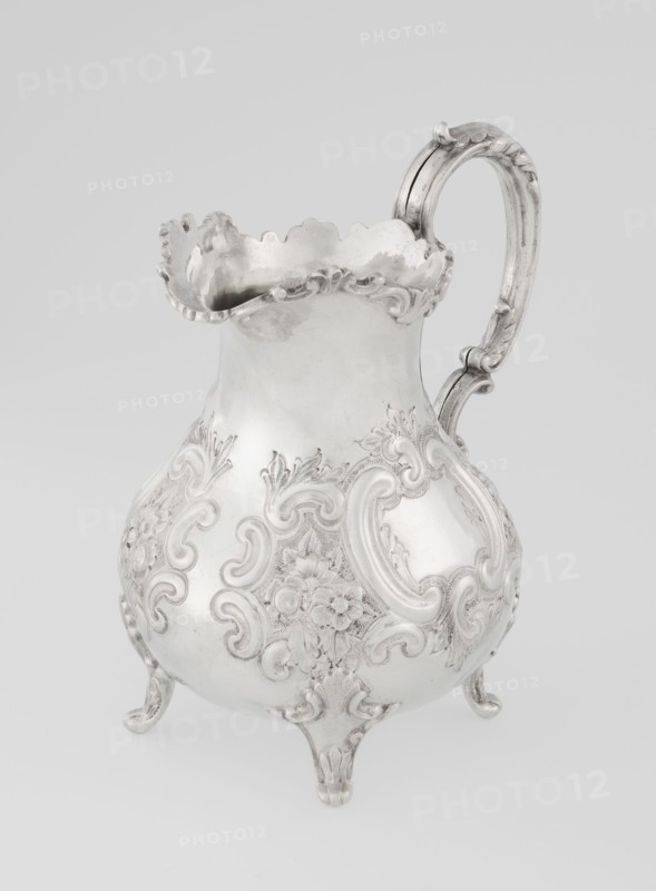 Milk jug by Mappin & Webb Ltd goldsmith, silversmith, dealer; fl. 1859–, neo-rococo style, pear-shaped milk jug supported on four volute legs, decorated with an acanthus leaf at the base and on the belly with rococo motifs in the form of channel-shaped jars, rocailles, flowers and acanthus leaves; volute handle with acanthus projections and a wide, raised spout with a serrated edge; dated 1895/1896; Sheffield, England, Great Britain; vessel; height 14.4 cm, width 9.5 cm, depth 8.5 cm; weight 266 g., A decorative metal pitcher with an ornate curved handle and a scalloped rim sits against a pale background; the pitcher has a bulbous body raised on a few molded feet and is covered in raised floral and scrollwork ornamentation, showing a shiny metallic surface in tones of silver and gray.