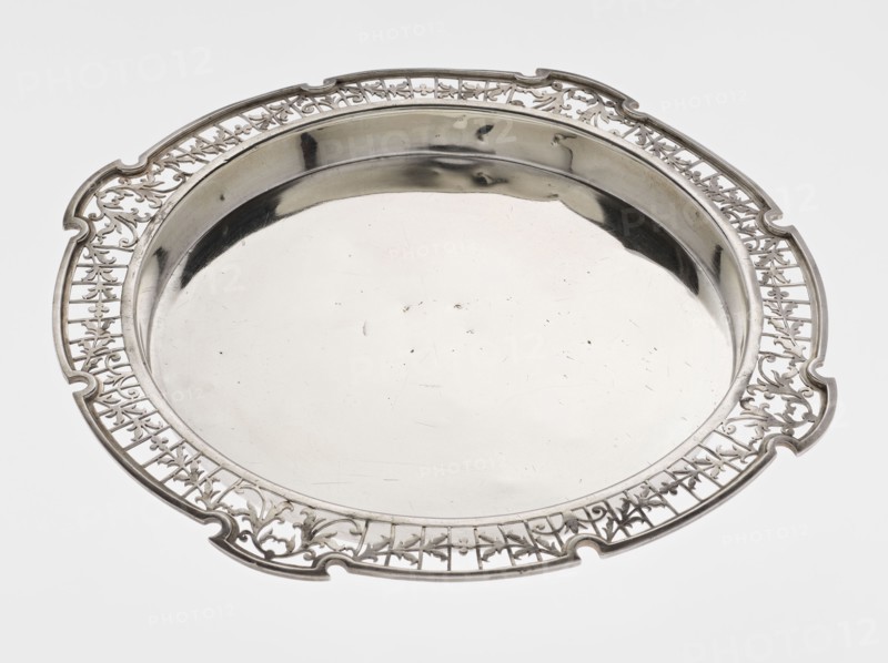Round tray, circular silver with smooth mirror and slightly raised, openwork collar decorated with plant tendril, irregularly cut edge bordered with profiled band; maker: Mappin & Webb Ltd goldsmith, silversmith, dealer; fl. 1859-; date: 1922/1923; place: Sheffield, England; function: table accessory; dimensions: height 1.8 cm, width 27.1 cm, depth 18 cm; weight: 576.5 g., A round metal tray with a reflective central surface and an ornate pierced rim is shown, primarily silver in color with highlights and darker reflections, set against a light gray background; the rim features an intricate openwork pattern with flowing, curving shapes and a few scalloped notches around the edge, while the central area is smooth and shiny with a few small surface marks.