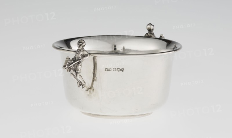 Porridge bowl porringer by Mappin & Webb Ltd goldsmith, silversmith, dealer; fl. 1859–. Cylindrical bowl with a flat bottom, slightly widening toward the top, with a turned-up edge edged by a smooth half-cylinder. Two side handles in the shape of figurines of boys dressed in jockey outfits, riding toy horses on sticks. Associated themes: jockeys, children's games, porridge. Material/function: vessel/porridge dish. Date: 1935–1936. Place: Sheffield, England, Great Britain. Dimensions: height 5.6 cm; width 16.3 cm; depth 10.5 cm; weight 125 g., A shiny silver bowl with a rounded rim and smooth curved sides sits against a light gray background, featuring a small sculpted figure attached to the side holding a drum and drumsticks and another small sculpted head peeking over the rim; the surface reflects highlights and shadows, showing metallic silver and soft gray tones with faint darker reflections.