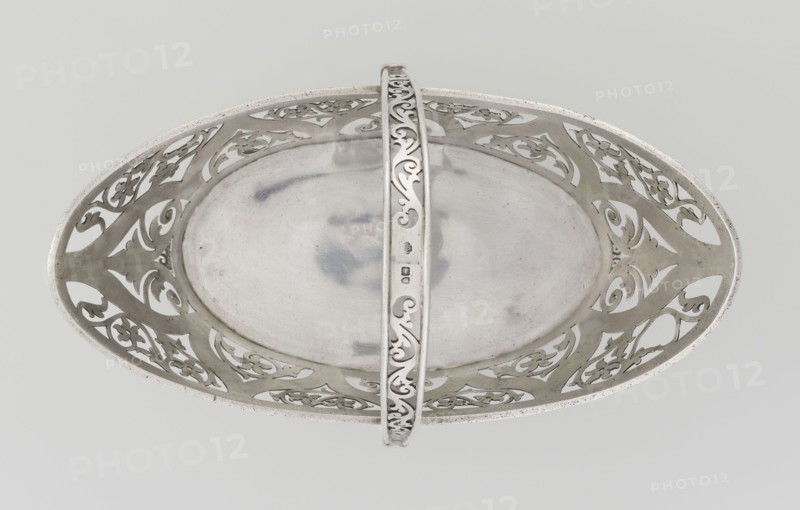 Bread Basket; maker: Mau, Hainrich fl. early 20th century maker; Hutton & Sons Ltd Sheffield, Birmingham, London; goldsmith and silversmith firm; fl. 1800–1930 maker; purchased provenance, scrap - purchased. Geometric and floral ornament; openwork; godron ornaments. Basket on four curved legs; oval, boat-shaped; sides decorated with openwork plant and floral twig motifs inscribed in an arched ogee back; ribbon reserves; ribbon upper edge; enclosed at the bottom by a godron half-cylinder. Handle in the form of a movable bow, openwork, decorated with plant tendrils. Date: 1901/1902, London, England, Great Britain. Vessel. Height 18 cm; width 22.7 cm; depth 3.7 cm; weight 388 g., An oval metal tray with a central plain recessed surface and an ornate pierced rim featuring repeating floral and scroll patterns, shown from above with a curved decorative handle crossing the center; the visible colors are silver, light gray, and subtle darker gray tones.