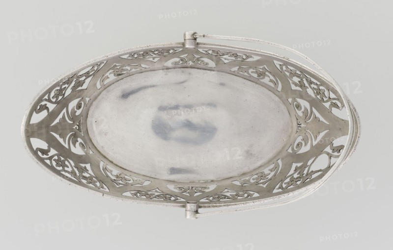 Bread basket by Hainrich Mau fl. early 20th century and Hutton & Sons Ltd goldsmith and silversmith firm, fl. 1800–1930. Oval, boat-shaped openwork silver basket on four curved legs, decorated on the sides with openwork plant and floral twig motifs set in an arched ogee back, ribbon reserves, ribbon upper edge, enclosed at the bottom by a godron half-cylinder. Handle is a movable bow in openwork decorated with plant tendrils. Geometric and floral ornament, godron ornaments. Dated 1901–1902, London, England, Great Britain. Vessel; height 18 cm, width 22.7 cm, depth 3.7 cm, weight 388 g., A decorative oval metal dish viewed from above with an openwork pierced border featuring ornate scroll and floral cutouts and a smooth central well showing subtle surface marks; the dish and its handles are metallic silver with areas of slightly darker gray patina against a pale gray background.