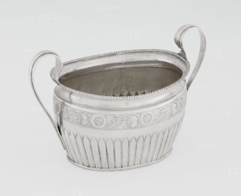 Sugar bowl by Hutton & Sons Ltd Sheffield, Birmingham, London; goldsmith and silversmith firm; active 1800–1930. Silver sugar bowl, oval with two ribbon handles; clasp vertically in the lower section. Decorated above with a band of stylised acanthus twig and an upper edge with a godron half-cylinder. Godron ornaments, plant twig motif. Function: sugar vessel. Inscription/markings: maker. Dated 1895–1896, London, England. Dimensions: height 11 cm, width 17.3 cm, depth 10.5 cm; weight 231 g., A metallic vessel with a silvery surface and a slightly reflective interior sits against a pale background; it has an oval opening, a decorative rim with a beaded edge, two tall curved handles rising from opposite sides, an engraved band of ornate swirling floral and circular motifs around its upper body, and a lower section with vertical fluted ridges, showing shades of silver, gray, and subtle warm reflections inside.