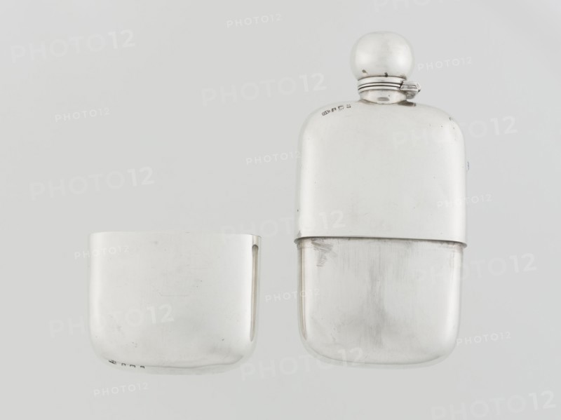 Hip flask, George & Sons Birmingham; goldsmith's firm; fl. 1825–1928. So-called cuboid hip flask with rounded corners; smooth, slightly curved; spout in the form of a narrow neck with an applied spherical hinged stopper; bottle narrowed in the lower part from the middle; on this part a slide-in container — a cup masking the narrowing — is placed; the interior of the cup and the spout are gilded. Alcohol vessel, 1907–1908. England, Birmingham. Height 15.5 cm, width 7.6 cm, depth 2.7 cm; weight 240 g., A photograph of a metallic hip flask and its matching cup, both shown against a pale gray background; the flask has a rounded rectangular shape with a smooth reflective surface and a spherical cap attached by a small hinged collar, and the cup is a shorter rounded rectangle with a similar smooth reflective finish; visible colors are silver, light gray, and subtle darker gray reflections across the surfaces.
