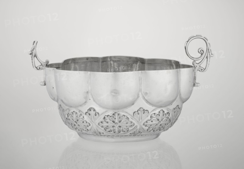 Porridge bowl porringer, Wakely & Wheeler Ltd London; goldsmiths; fl. 1791–1958, hemispherical bowl decorated with relief acanthus rosettes in the lower part, knobbed above, octagonal-shaped rim; two volute handles on the sides one half-preserved. Materials/technique: silver implied. Function: porridge dish. Ornament: rosette, acanthus, relief decoration. Date: 1891/1892. Place: London, England, Great Britain. Dimensions: height 7.6 cm; width 24 cm; depth 19.5 cm; weight 415 g., A decorative metal bowl with ornate handles sits centered on a smooth surface, showing a reflective finish in tones of silver and gray; the bowl has a scalloped rim and a rounded body with embossed floral-like patterns along the lower portion, and a subtle reflection of the bowl appears on the surface beneath it.