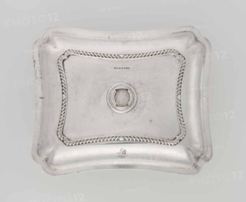 Four-sided platter by Bradbury, Thomas & Sons Sheffield; goldsmiths; fl. ca. 1769–1943. Four-sided platter with slightly concave, raised sides; upturned, irregularly cut rim, decorated with a band of godron ornament. Engraved on the side with a rooster standing on a crown. Dated 1903/1904, London, England, Great Britain. Dish. Height 3.9 cm; width 25 cm; depth 20.8 cm; weight 684 g., A rectangular metallic lid with rounded, scalloped corners and an indented central area, featuring a raised circular opening at the center and a decorative twisted rope-like border framing the inner panel; the surface shows subtle scratches and faint tarnish spots. The visible colors are silver, light gray, and darker gray tones in the worn areas.