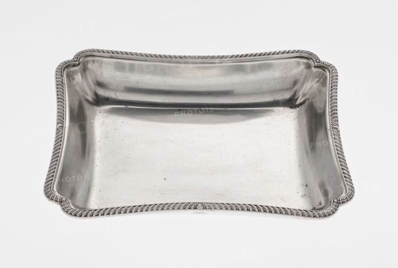 Four-sided platter by Thomas Bradbury & Sons Sheffield; goldsmiths; fl. ca. 1769–1943. Four-sided platter with slightly concave, raised sides; upturned, irregularly cut rim, decorated with a band of godron ornament. Engraved on the side depicting a rooster standing on a crown. Dated 1903/1904, London, England. Dish. Height 3.9 cm; width 25 cm; depth 20.8 cm; weight 684 g., A rectangular shallow tray with rounded corners and a raised rim featuring a twisted rope-like border, the tray surface and sides are smooth and reflective with visible scuffs and small blemishes, and the overall colors visible are metallic silver with highlights and darker gray reflections against a white background.